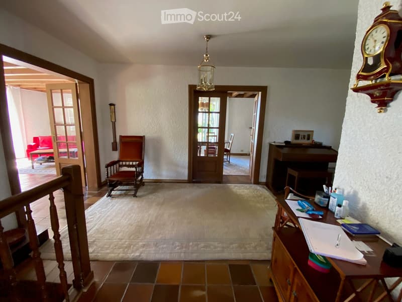 Spacious hallway with tiled flooring, wooden staircase, a rocking chair, a small table, a desk with office supplies, and a piano in the background.