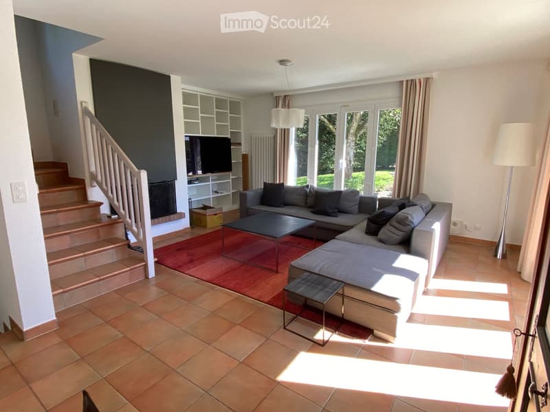 Bright living room with a grey sectional sofa, coffee table, small stool, TV on a shelf, and a staircase leading upstairs.