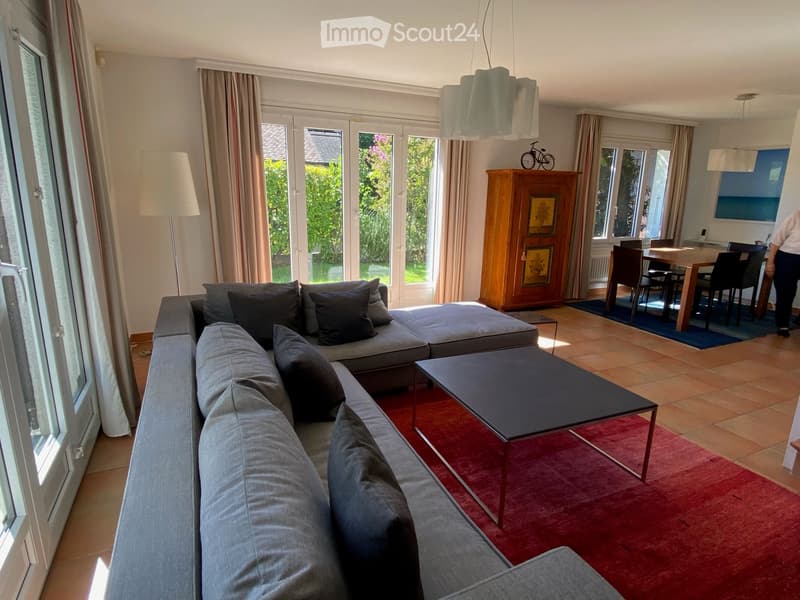 Modern living room with large windows, grey sectional sofa, coffee table, dining area with table and chairs, and a wooden cabinet with artwork.