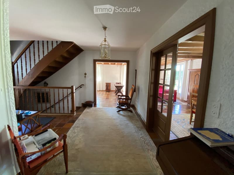 Spacious hallway with wooden staircase, chandelier, and open doors leading to other rooms. Visible furniture includes a rocking chair, small table, and books on a side table.