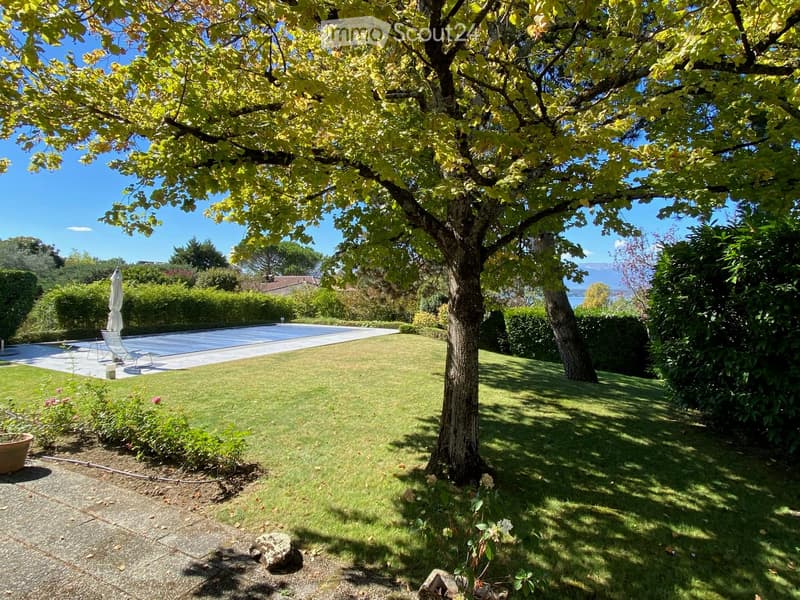 Well-maintained garden with a large tree, neatly trimmed grass, a swimming pool, lounge chairs, and a view of the surrounding landscape.