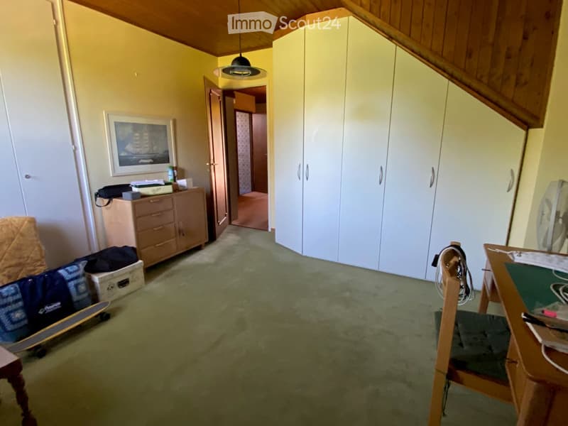 A furnished room with a slanted wooden ceiling, carpeted floor, and several pieces of furniture including a chair, table, cabinet, and fan. A skateboard and a picture frame are also visible.