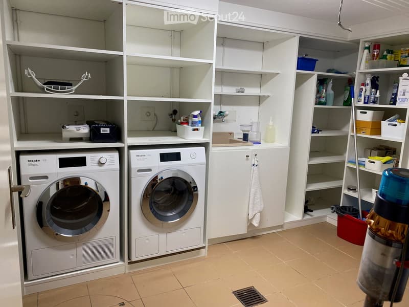 A modern bathroom with white cabinets, a washing machine, a dishwasher, shelves filled with cleaning supplies, and a towel hanging on the side.