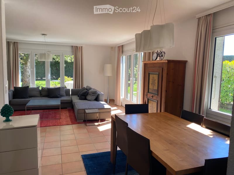 Bright living and dining area with large windows and sliding doors leading to a garden. Features a grey sectional sofa, coffee table, dining table with chairs, and a wooden cabinet with a clock on top.