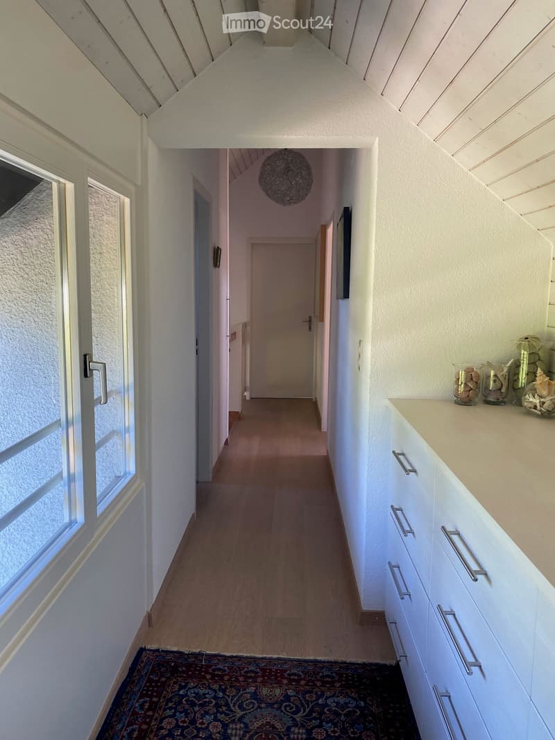 A narrow hallway with wooden flooring, white walls, and a hanging spherical light fixture. A small cabinet with glass jars and decorative items is visible on the right side.