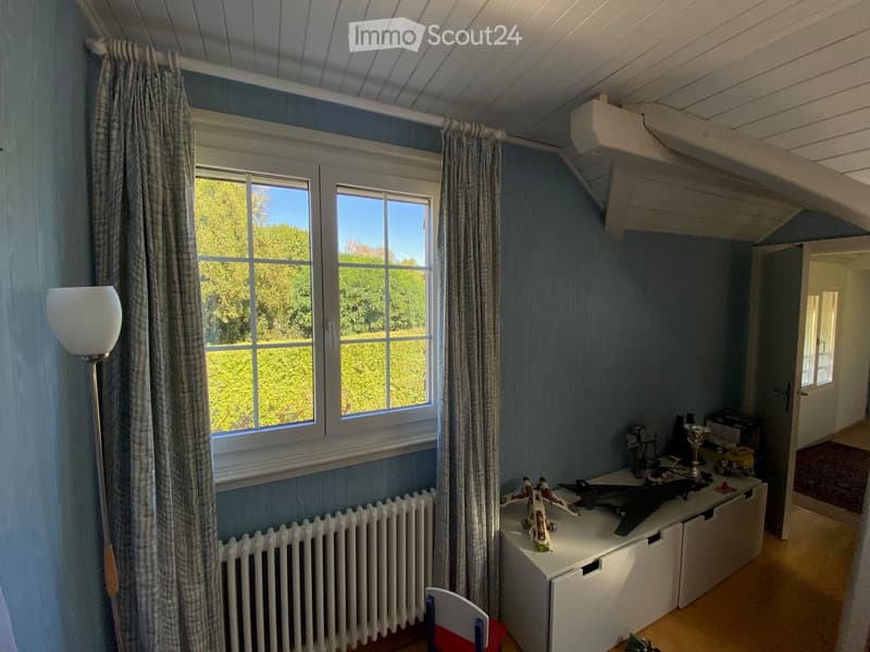 A cozy room with light blue walls, wooden floor, large window with curtains, a white radiator, a white table with model airplanes, a red and white chair, and a floor lamp.