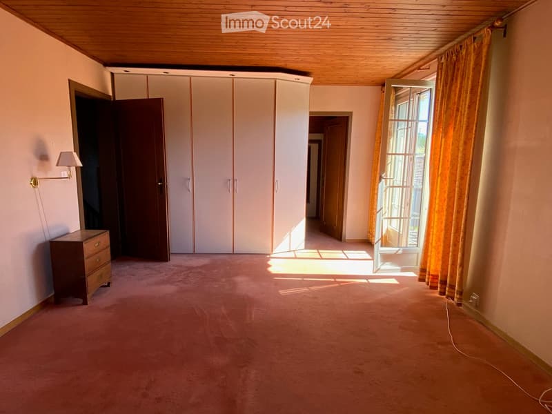 A spacious, well-lit hallway with carpeted flooring, a wooden ceiling, and large windows with yellow curtains. Features include a built-in white cabinet, a wall-mounted lamp, and a small wooden drawer unit.