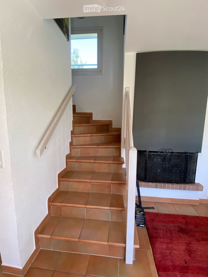 Staircase with beige tiles, white walls, and a small window at the top. A red rug is placed on the floor near the bottom of the stairs.