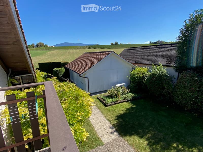 Balcony overlooking a well-maintained garden with a small shed, lush greenery, and a scenic view of vineyards and mountains in the distance.