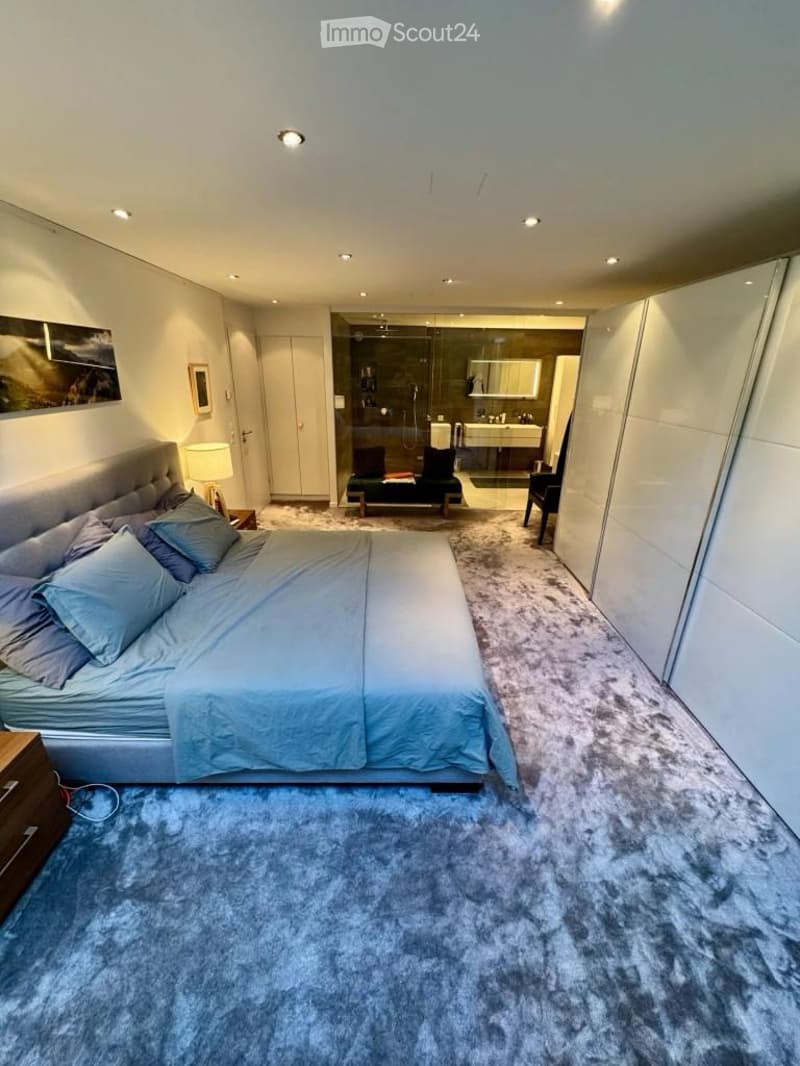 Modern bedroom with a large bed, nightstand, lamp, and a walk-in closet with a mirror. The room features a plush blue carpet and recessed lighting.