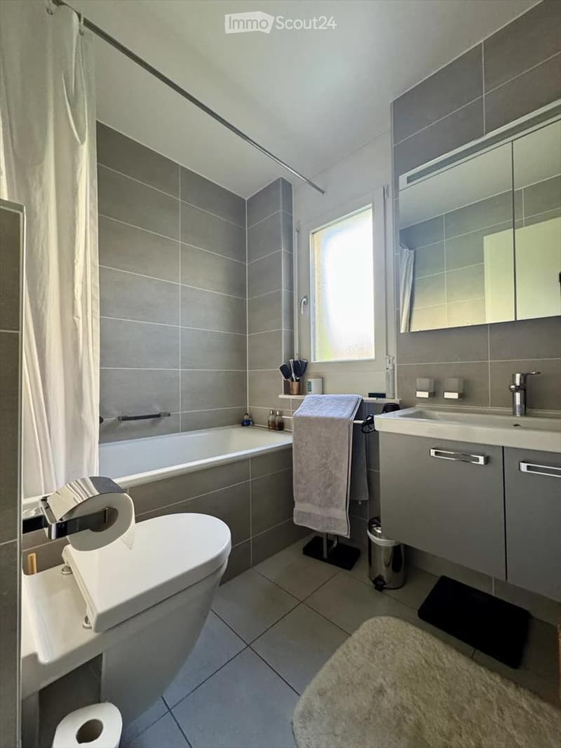 Modern bathroom with grey tiles, a bathtub, toilet, sink, mirror, and a towel rack with a towel. The space includes a window and a small shelf with toiletries.