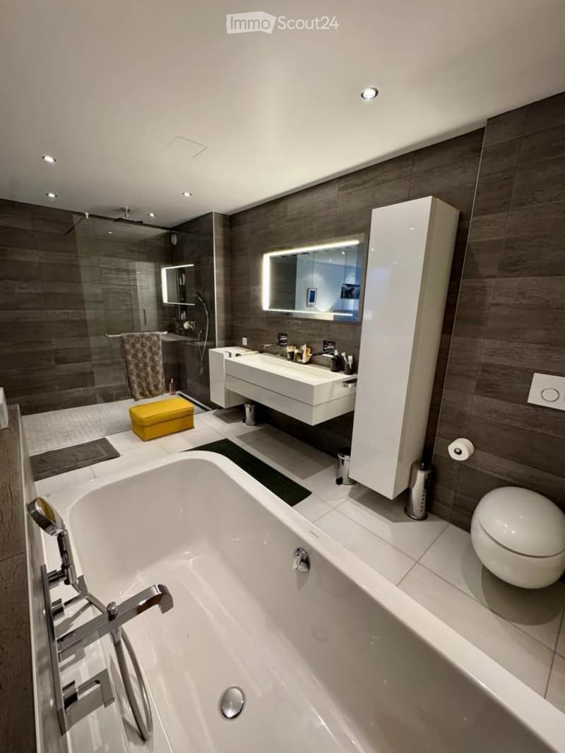 Modern bathroom with a large bathtub, toilet, sink, illuminated mirror, and a yellow stool. The walls are clad in dark wood, and the floor is tiled.
