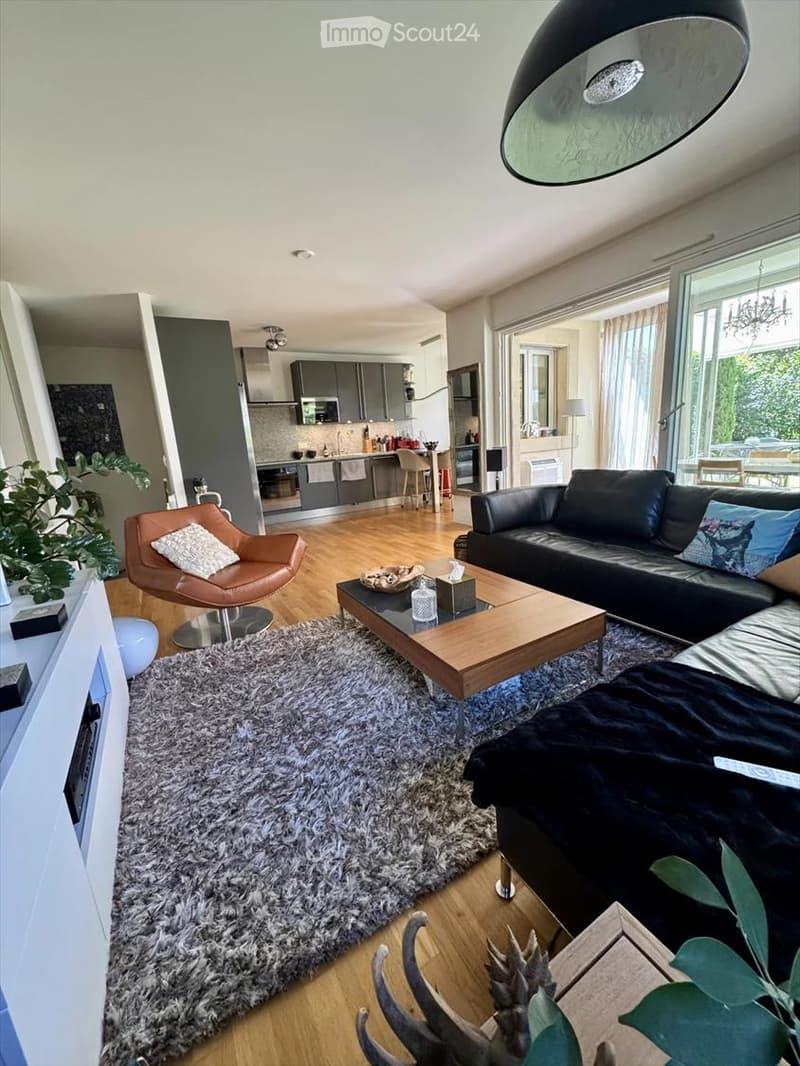 Modern living room with a black leather sofa, wooden coffee table, shaggy rug, and a leather armchair. The space features a sleek kitchen area with grey cabinets and stainless steel appliances visible in the background.