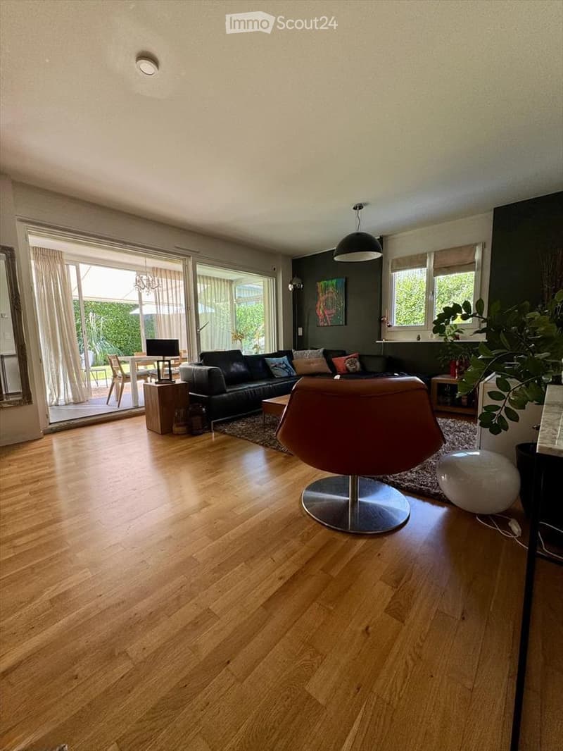 Modern living room with hardwood flooring, large windows, a black leather sofa, a brown leather chair, a coffee table, a dining area with a table and chairs, and a mirror on the wall.