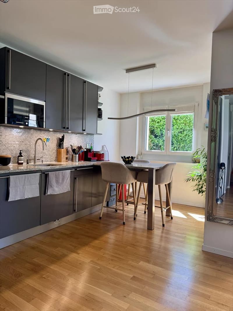 Modern kitchen with dark grey cabinets, wooden flooring, and a central island with bar stools. Features a sink, microwave, and various kitchen items on the counter.