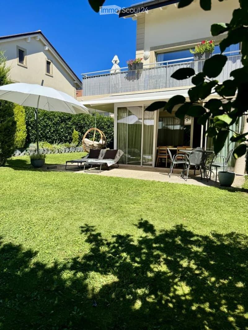 Well-maintained garden with a lawn, patio seating area, and a large umbrella. The patio includes lounge chairs, a dining table, and chairs, with a modern house featuring a balcony in the background.