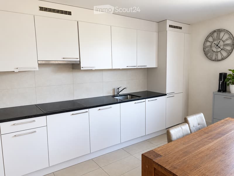 Modern kitchen with white cabinets, black countertops, induction cooktop, sink, and a wooden dining table with chairs. A wall-mounted clock and a coffee maker are also visible.