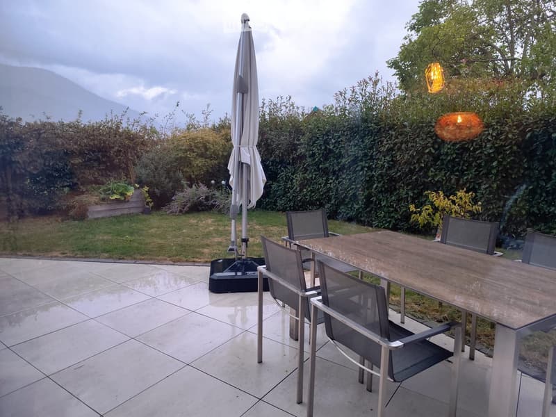 Outdoor patio with tiled floor, wooden table, metal chairs, umbrella, and surrounding greenery.