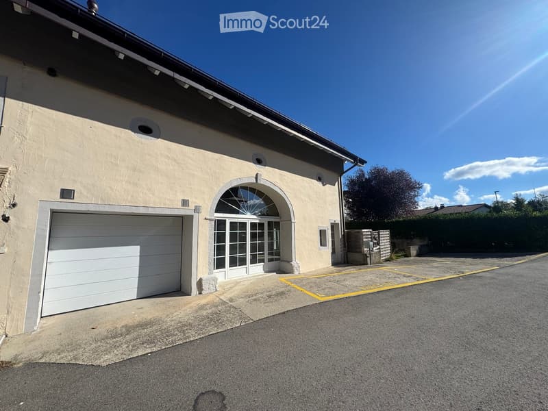 Single-story building with a garage and arched entrance, surrounded by a paved area with marked parking spaces.