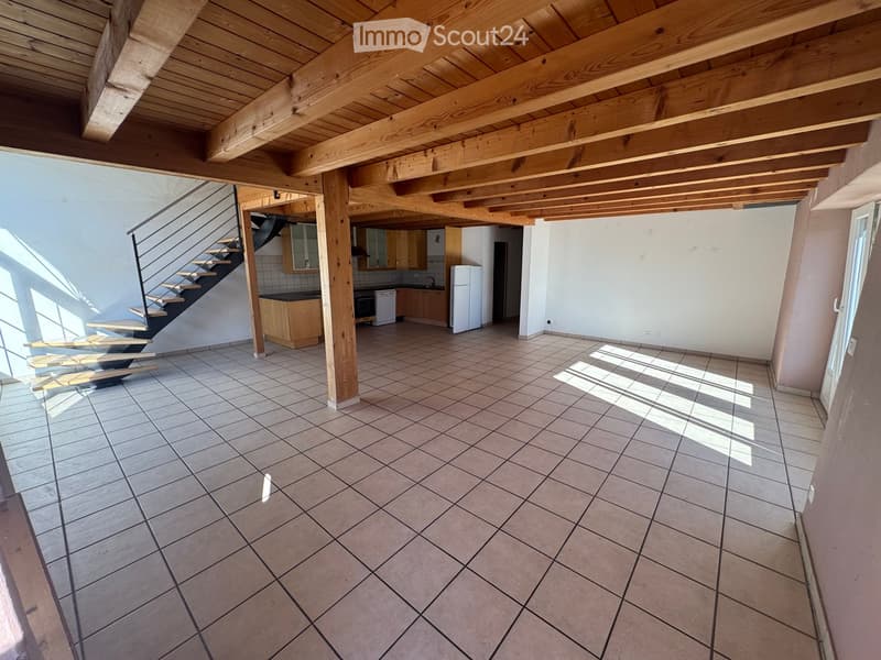 Empty room with tiled floor, wooden ceiling beams, staircase leading to another level, and a small kitchen area with cabinets and appliances.