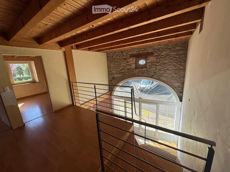 Spacious hallway with wooden flooring, exposed wooden beams on the ceiling, and a modern metal railing leading to a lower level. Features a large arched window and a smaller window on the left side.