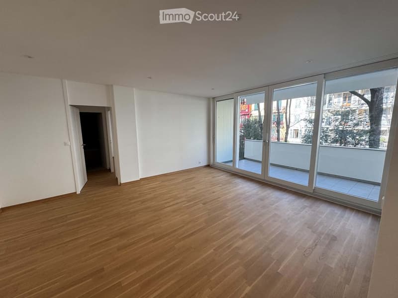Empty room with wooden flooring, white walls, large sliding glass doors leading to a balcony, and an open doorway to another room.