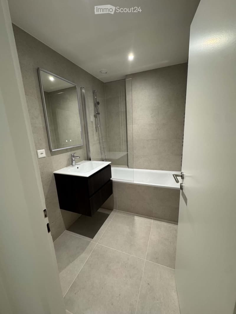 Modern bathroom with a white sink, dark wooden cabinet, bathtub, and large mirror. Light-colored tiles on walls and floor.