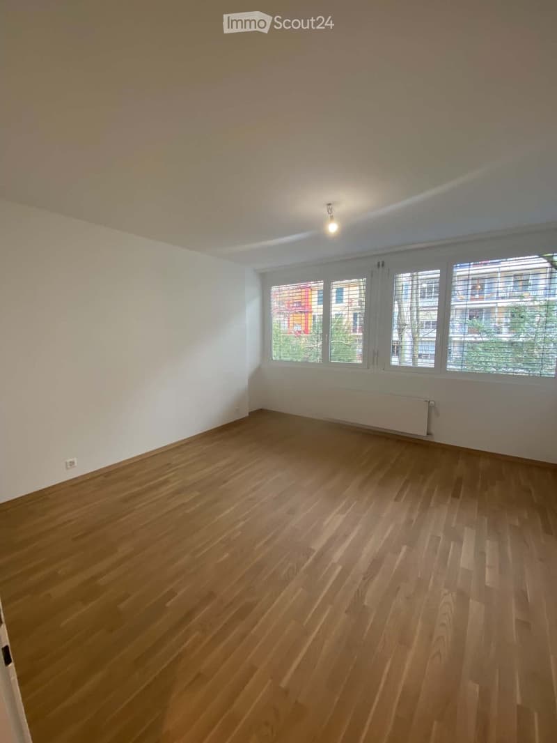 Empty room with wooden flooring, white walls, large windows with blinds, and a radiator under the windows.