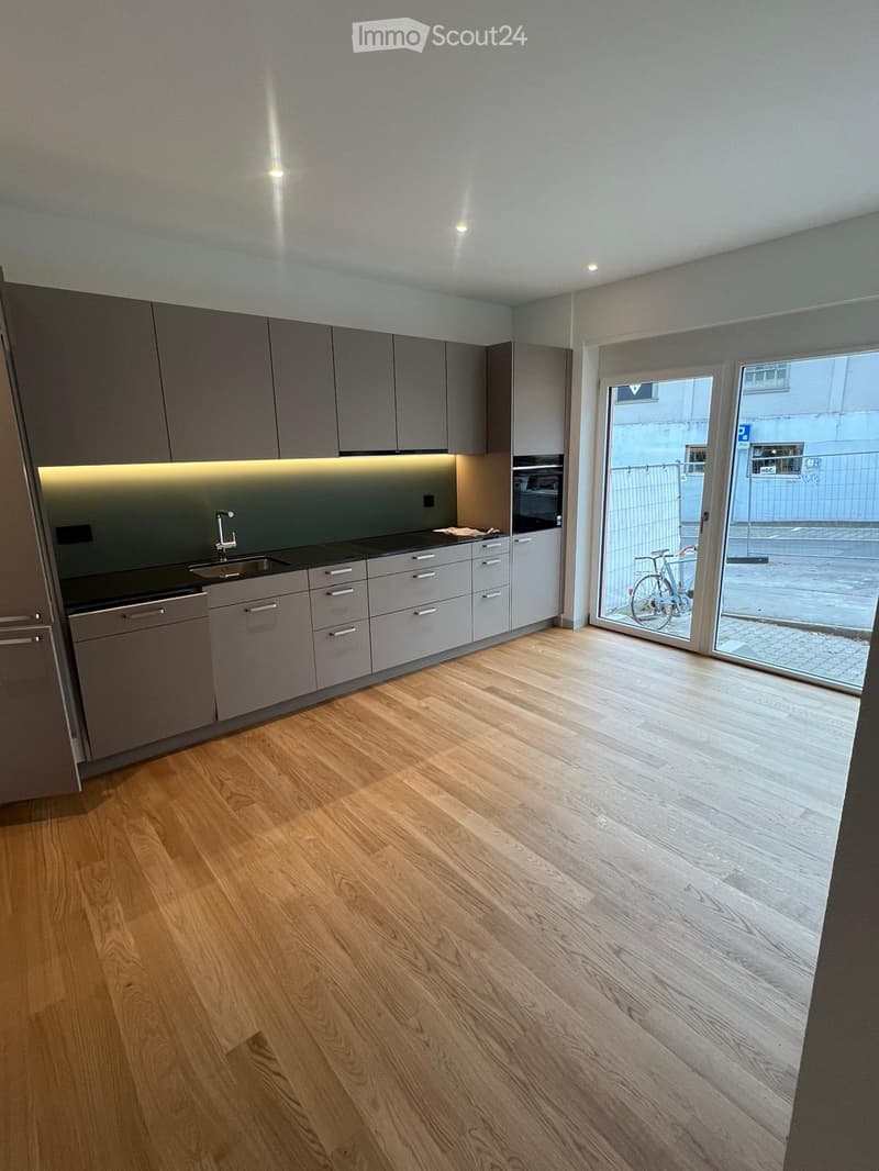Modern kitchen with grey cabinets, wooden flooring, and built-in appliances including a sink, oven, and dishwasher. A large sliding glass door leads to an outdoor area.