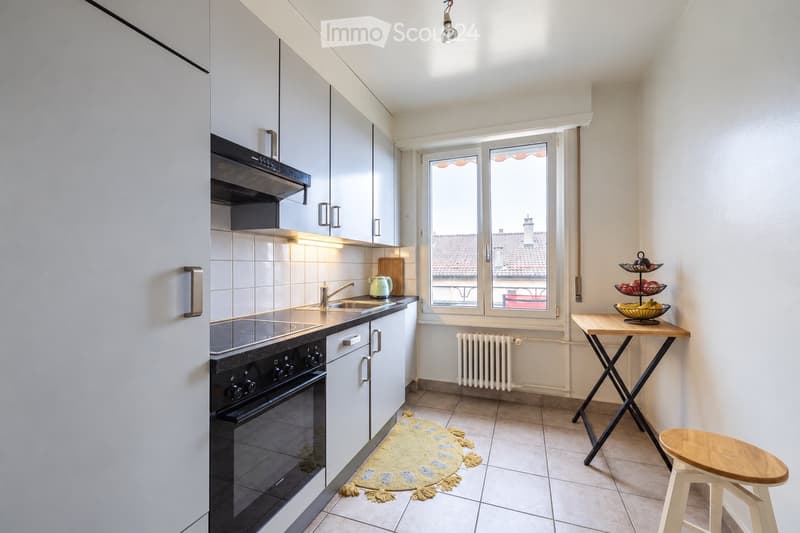 Modern kitchen with white cabinets, stainless steel appliances, a window overlooking a tiled roof, a small table with a fruit basket, and a stool.