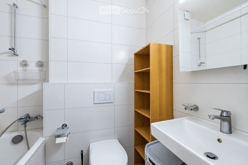 Modern bathroom with white tiled walls, a bathtub, toilet, sink, mirror, and a wooden shelf for storage.