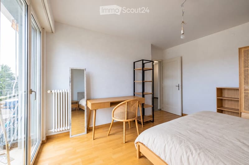 Modern bedroom with light wooden flooring, a bed with white bedding, a wooden desk with a woven chair, a tall mirror, and a black shelving unit.