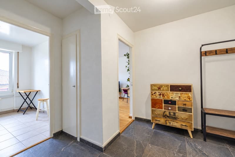 A small, well-lit hallway with tiled flooring and white walls. A small table and stool are positioned near a window, while a colorful, patterned cabinet and a wooden shelf are visible on the right side. The hallway leads to another room with a chair and a