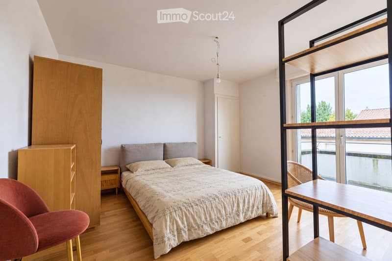 Modern bedroom with a wooden bed, grey headboard, patterned bedspread, pink armchair, wooden table, and a large window with a view of the outside.