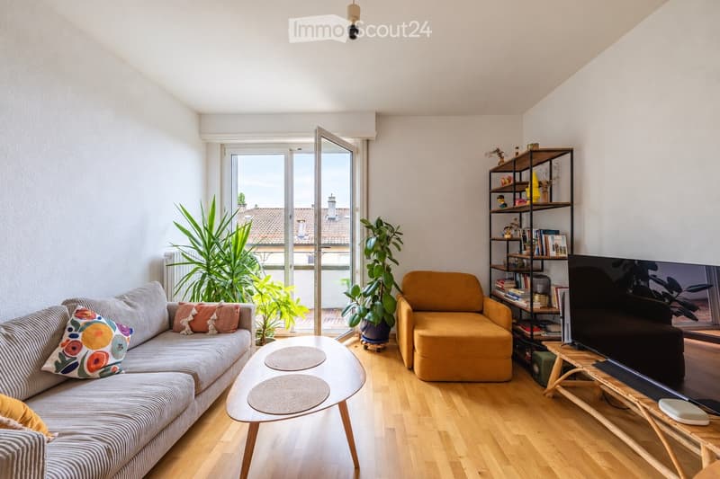 Modern living room with wooden flooring, large windows opening to a balcony, grey sofa with colorful cushions, yellow armchair, coffee table, bookshelf, and television.