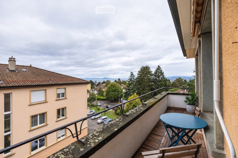 Balcony with wooden decking, metal railing, small round table, chairs, and potted plant. Offers a view of a residential area with houses, trees, and a road.