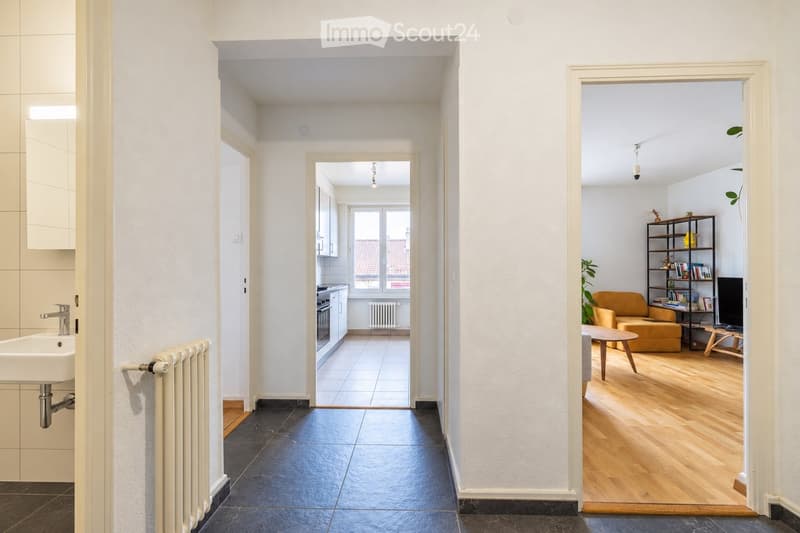 A modern, well-lit hallway with white walls and dark tiled flooring, leading to a kitchen and a living room area. The kitchen features white cabinets, a sink, and a radiator, while the living room includes a yellow armchair, a coffee table, a bookshelf, a