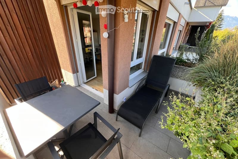 Balcony with a table, chairs, and a lounge chair, surrounded by plants and a view of the mountains.