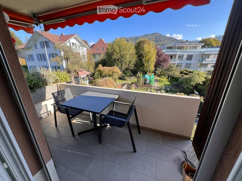 Balcony with a table and chairs, overlooking a scenic view of houses, trees, and mountains.