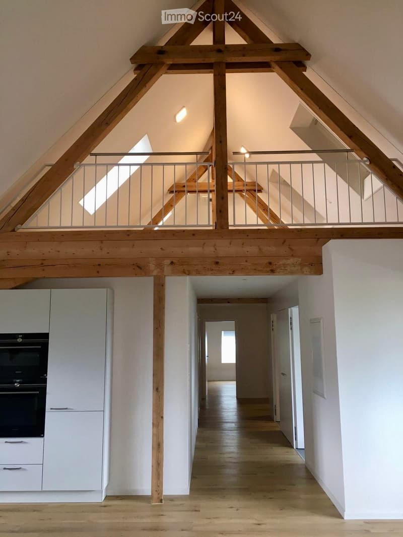 Bright, open hallway with wooden floors and white walls, leading to a kitchen area with white cabinets and a built-in oven. The hallway features exposed wooden beams and a metal railing leading to an upper level.