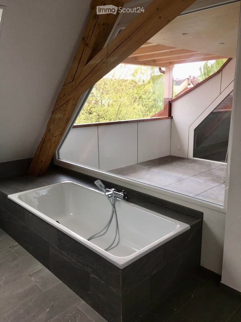 Modern bathroom with a large bathtub, sleek faucet, dark tiled walls and floor, and a large window offering a view of greenery.