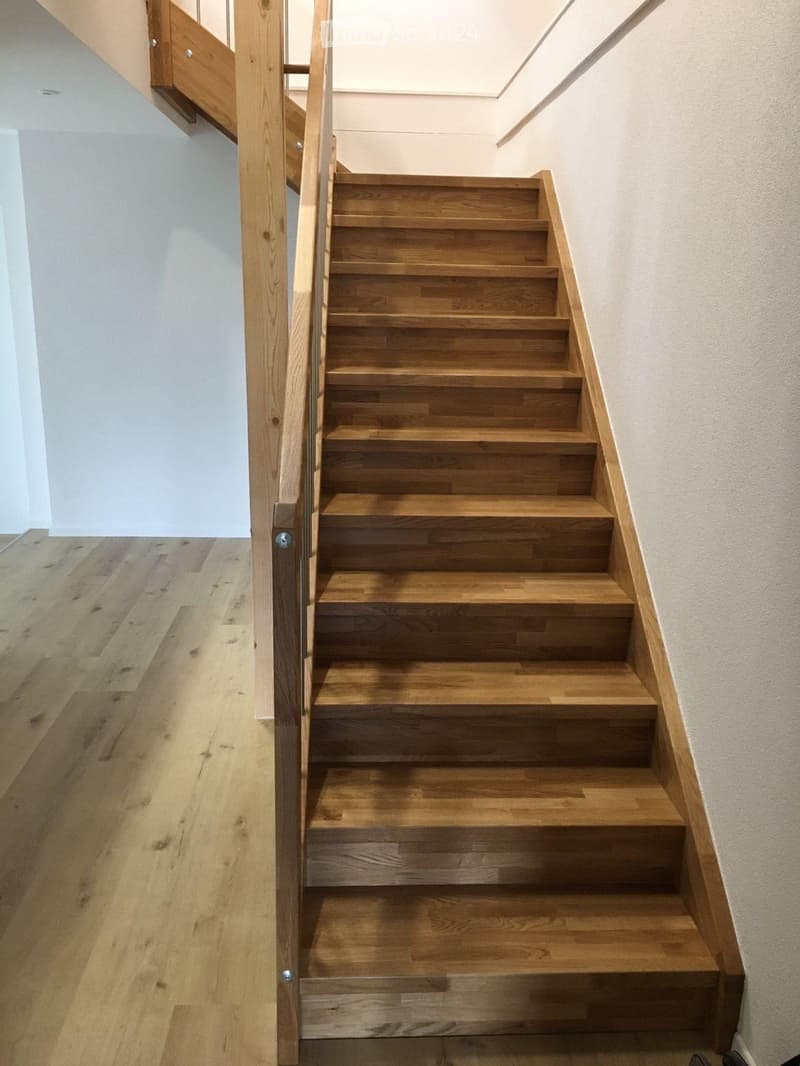 Wooden staircase with a modern glass railing, light-colored wooden flooring, and white walls.