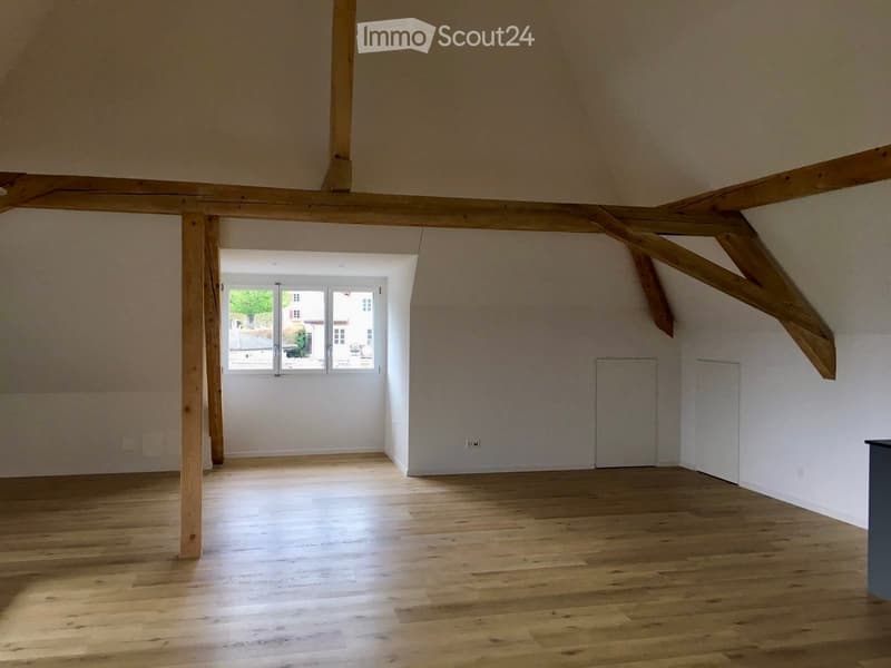 Empty room with wooden flooring, exposed wooden beams on the ceiling, and a large window letting in natural light.