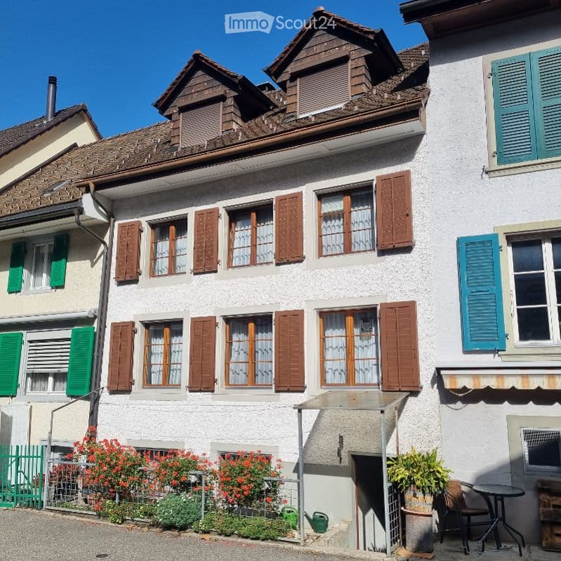Traditional Swiss house with white plaster walls, wooden shutters, multiple windows, and a small garden area with flowers and a table.