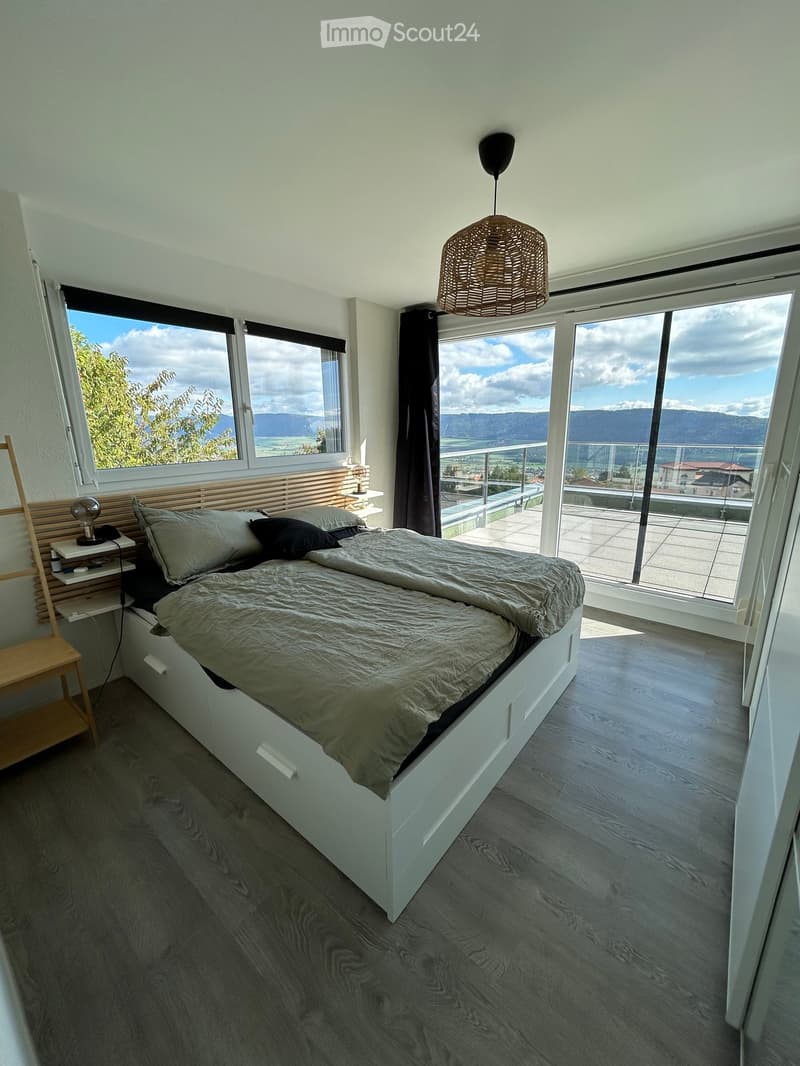 Modern bedroom with large windows offering a scenic mountain view, a white bed with grey bedding, a nightstand with a lamp, and a pendant light hanging from the ceiling.