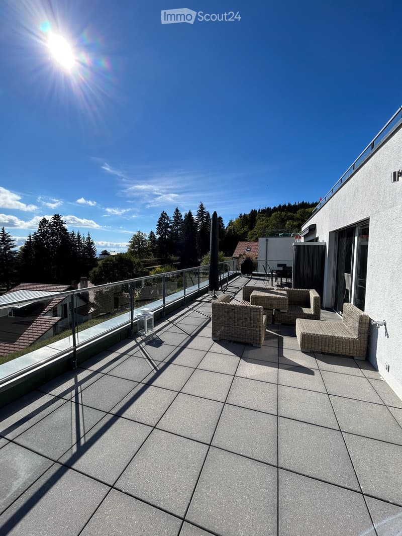 Balcony with modern furniture, including a sofa, table, and chairs, overlooking a scenic view of trees and houses.