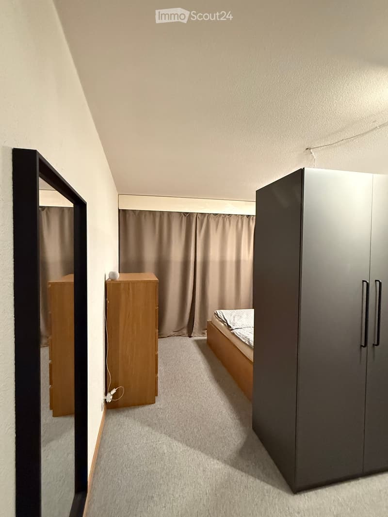 Cozy bedroom with a bed, wardrobe, nightstand, and mirror. The room features light-colored walls, carpeted flooring, and a slanted ceiling.