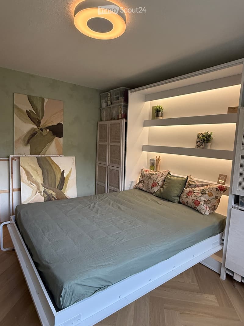 Modern bedroom with a raised bed, built-in storage, and ambient lighting. Features include a grey duvet, floral pillows, a white cabinet, and decorative shelves with small plants and artwork.