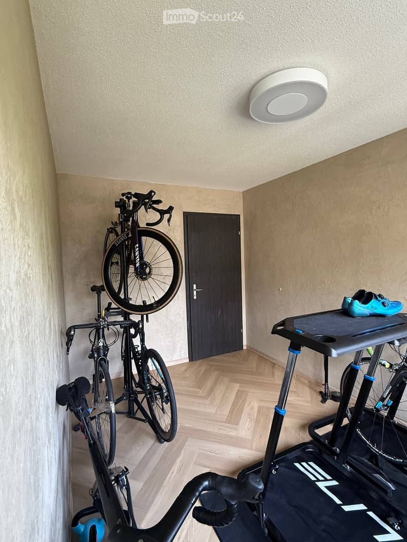 Exercise room with wooden flooring, a mounted bicycle, an exercise bike, and a pair of blue shoes on the bike's console.