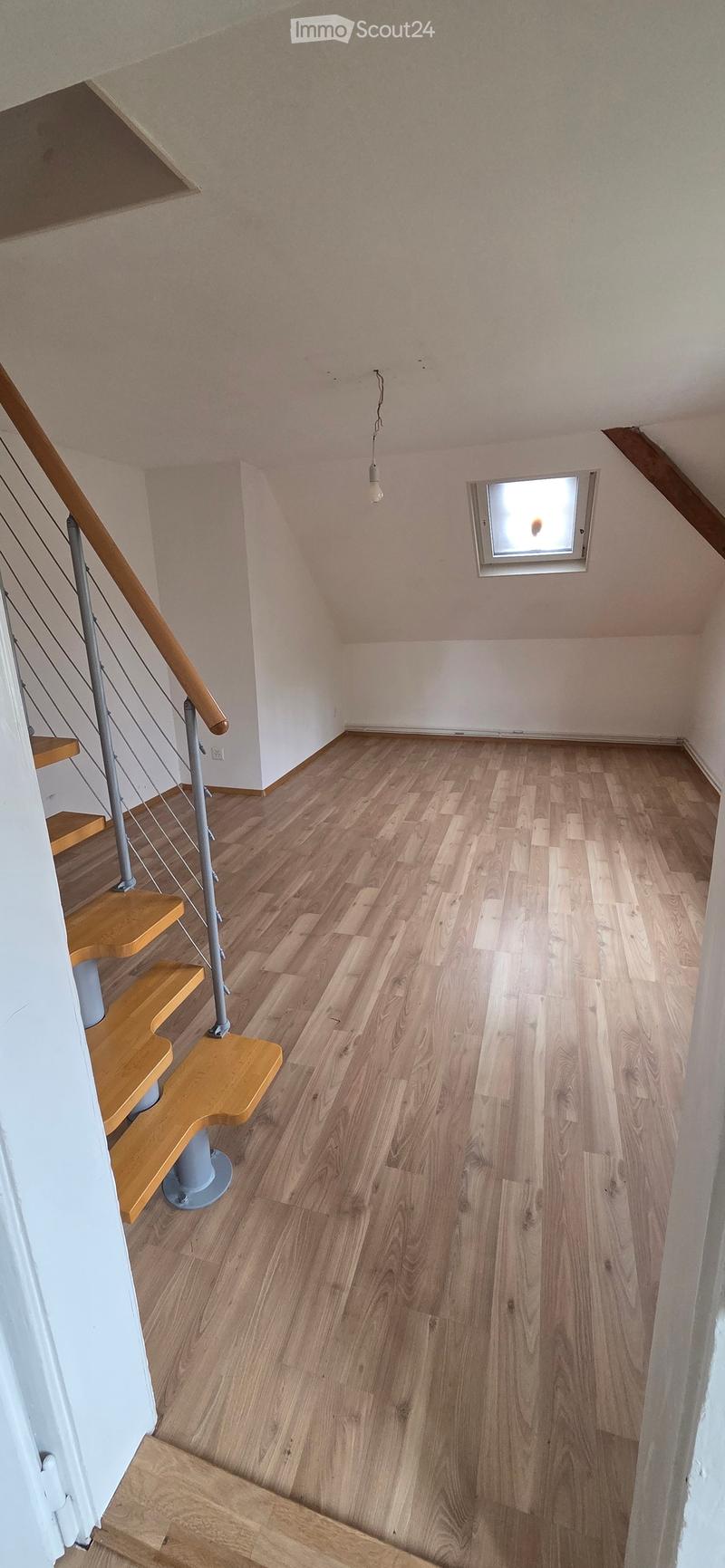 Empty hallway with wooden flooring, white walls, and a staircase leading upwards. A small window is visible on the wall.